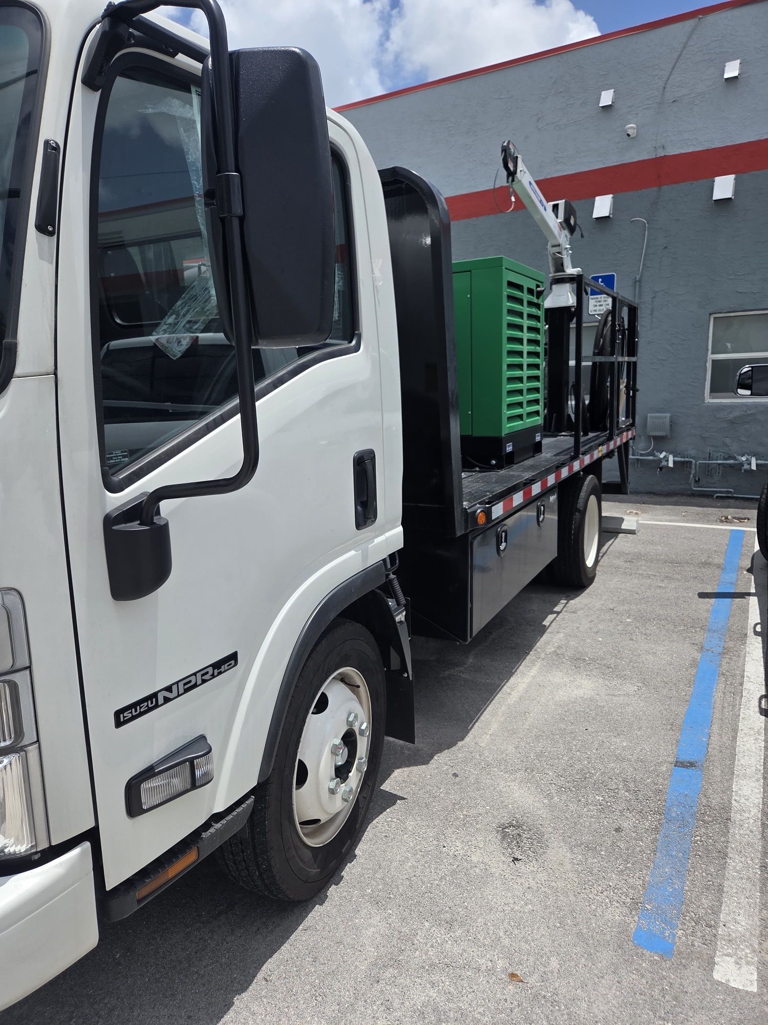 White truck with black flatbed; green equipment and crane visible on the bed. Parked next to a building.
