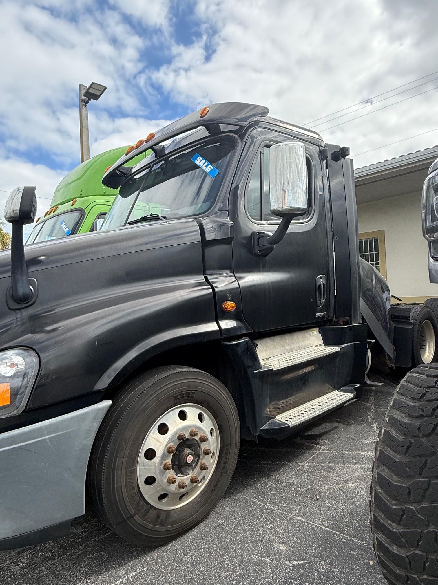 Black semi-truck parked outdoors on a sunny day. The truck has a gray bumper and a light blue tag on the window.