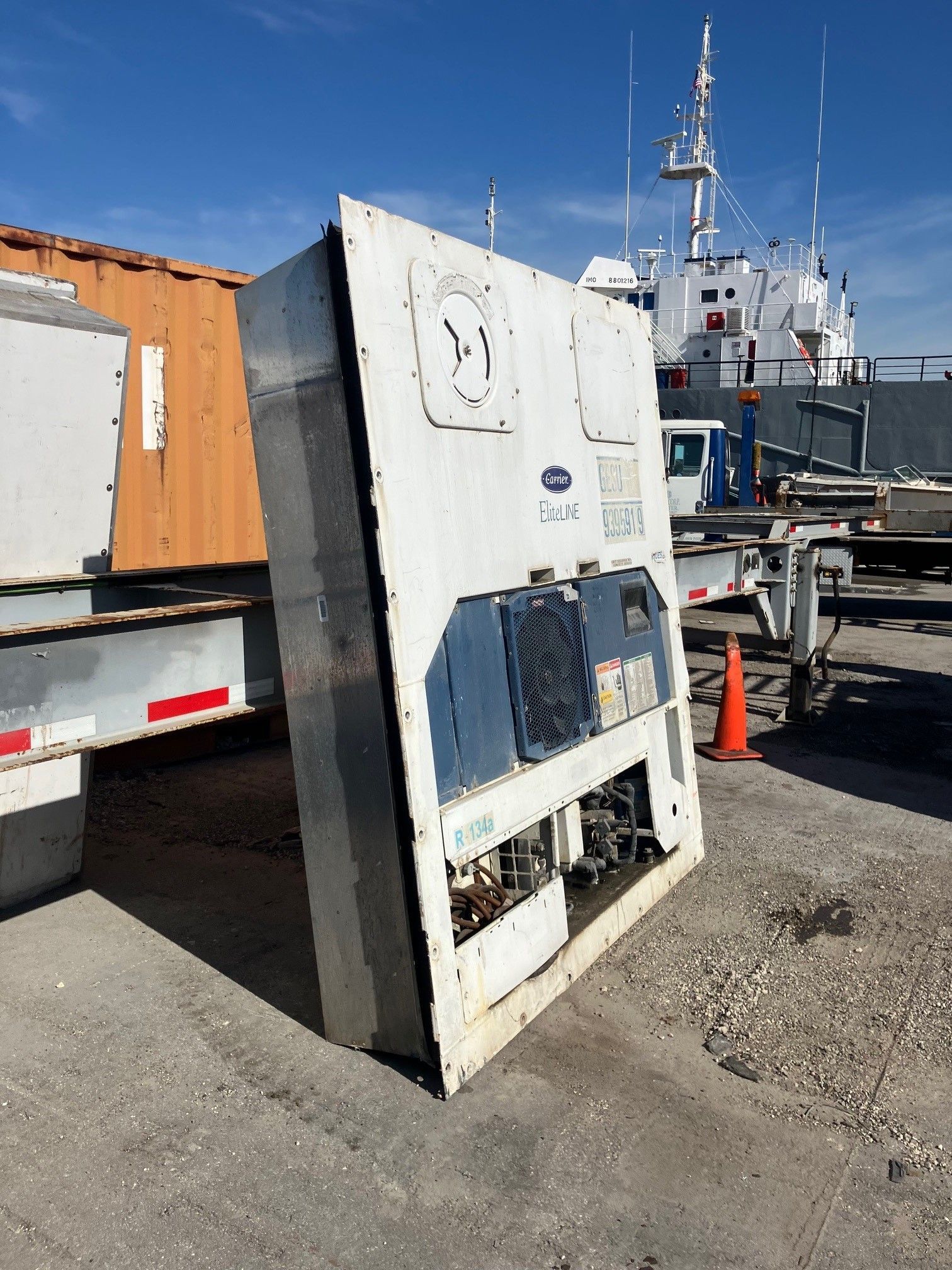Large, white refrigeration unit leaning on a dock, with a ship in the background.
