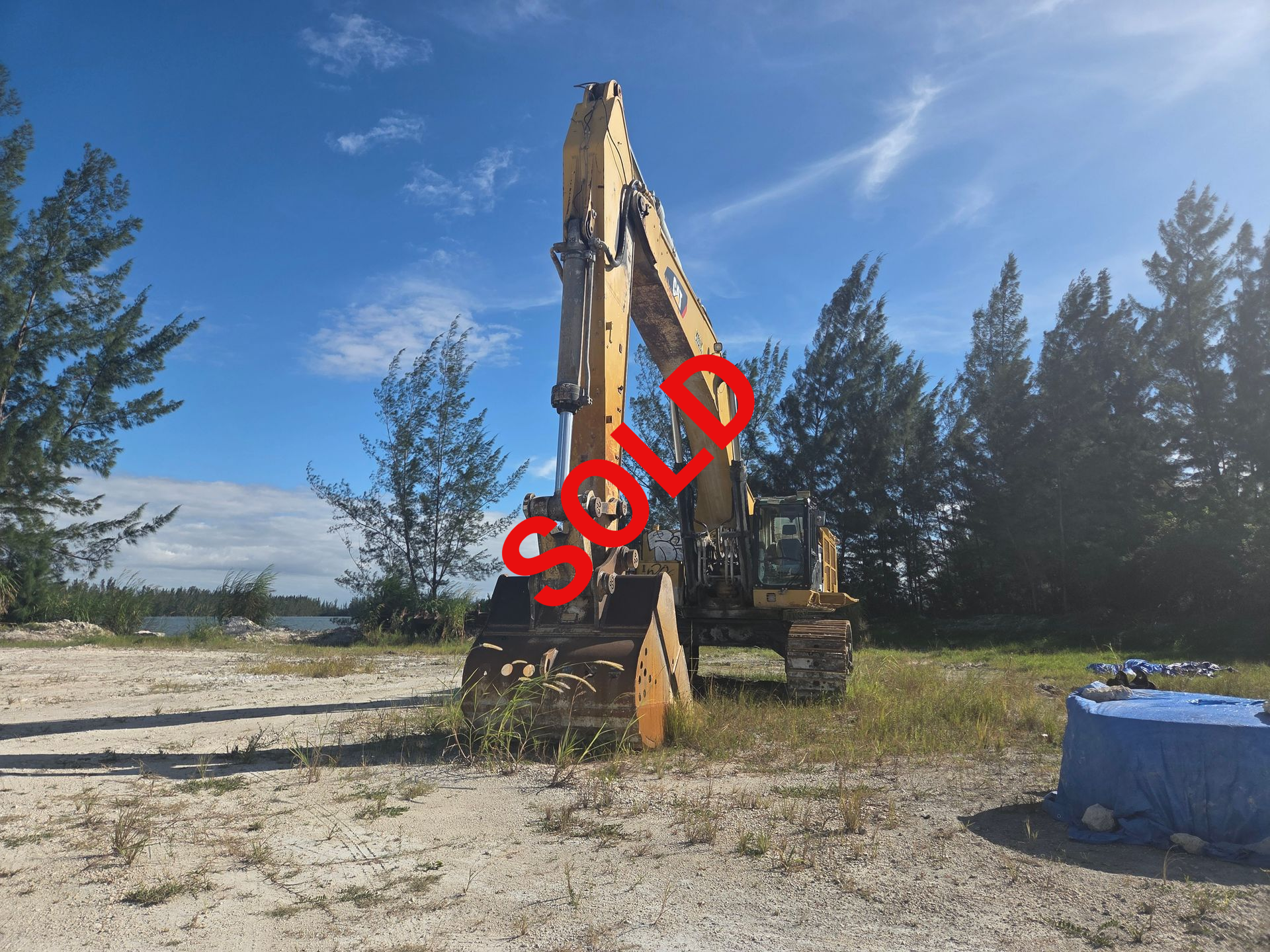 Yellow excavator on a dirt ground under a blue sky near trees and water.