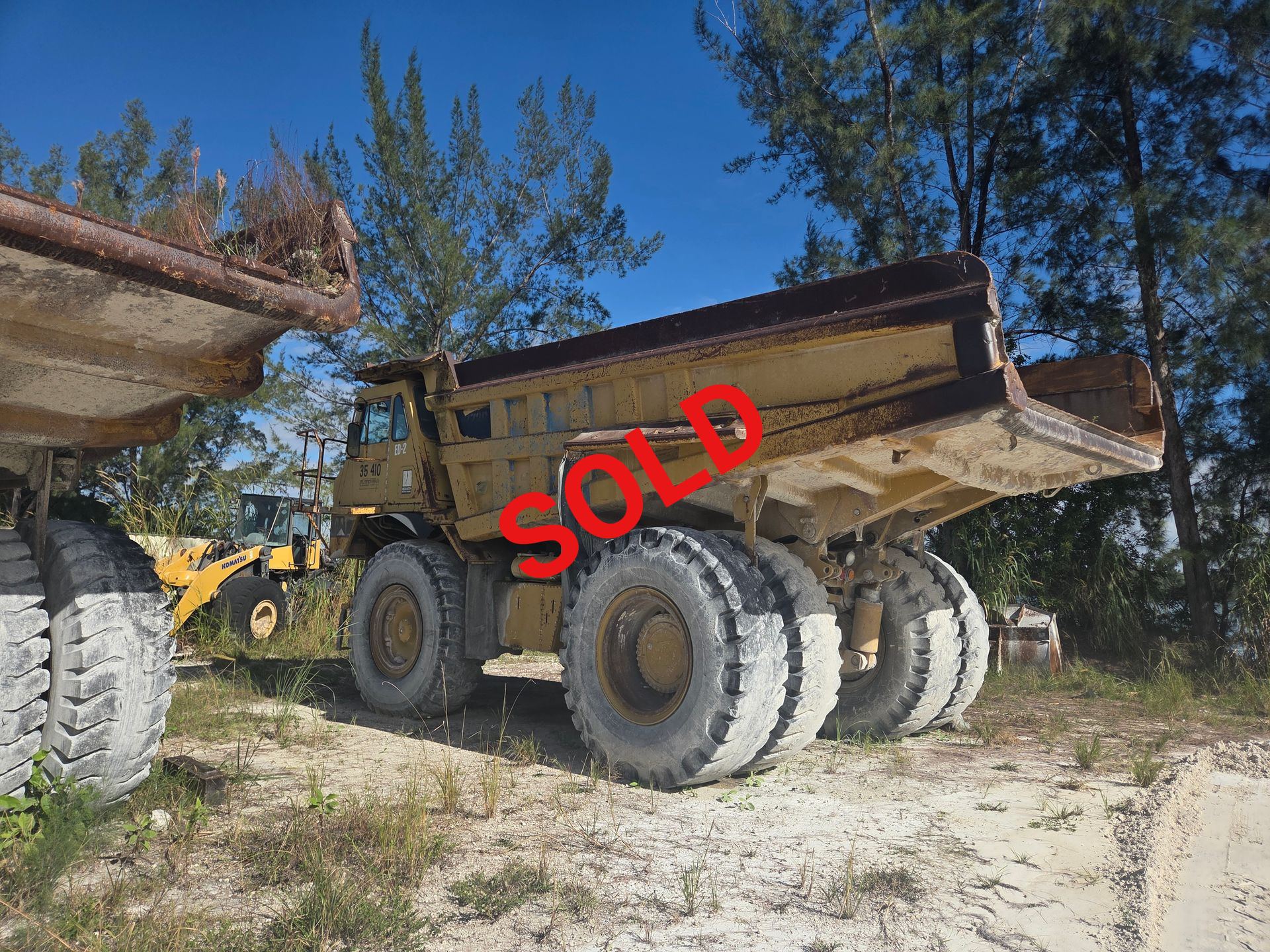 Yellow dump truck with large tires in an outdoor setting. Another truck and trees are visible.