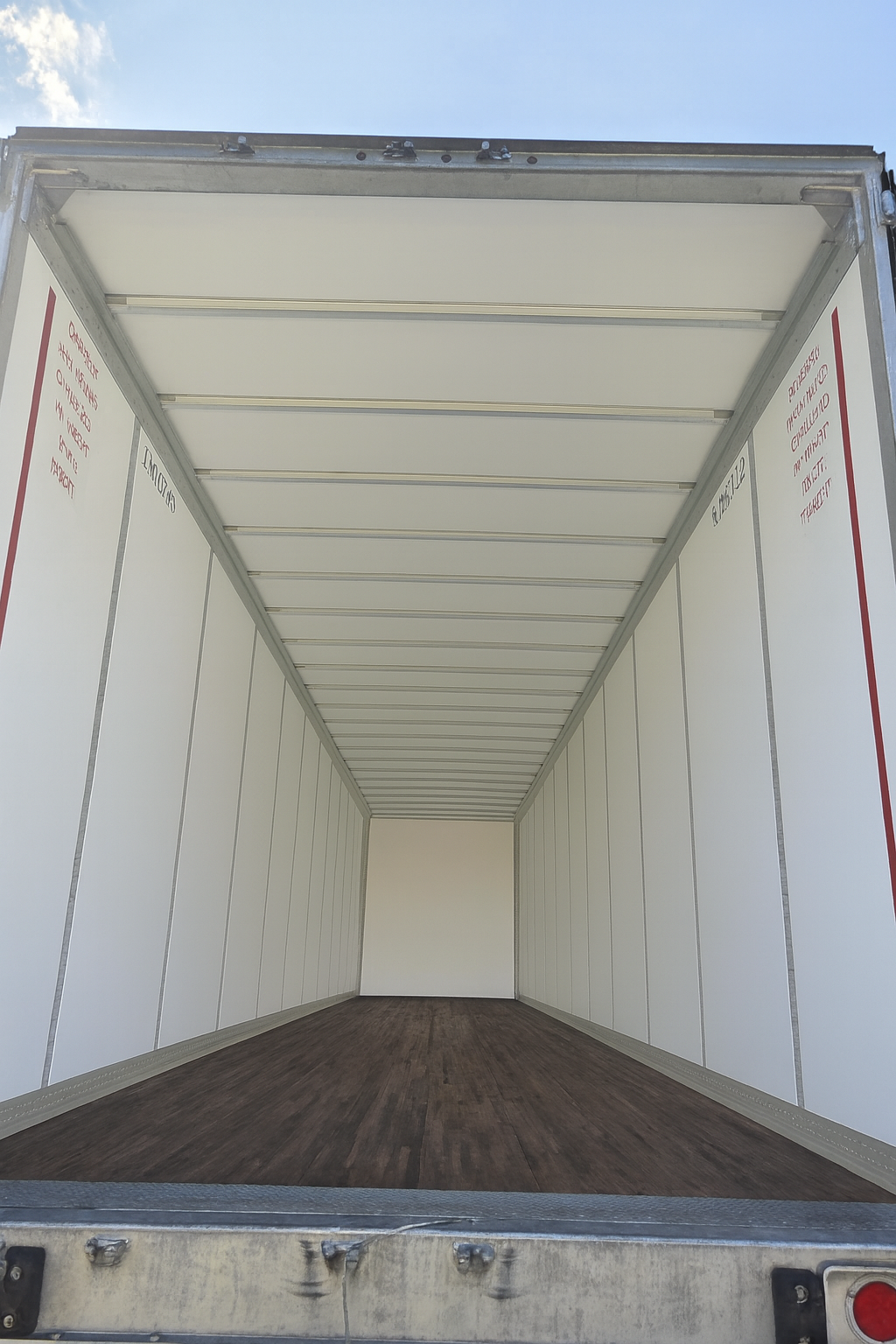 Empty interior of a semi-trailer. White walls and ceiling, dark brown floor, and a bright sky is visible at the opening.