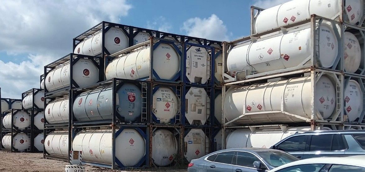 White cylindrical tanks in metal frames stacked outdoors. Blue sky, and car below.