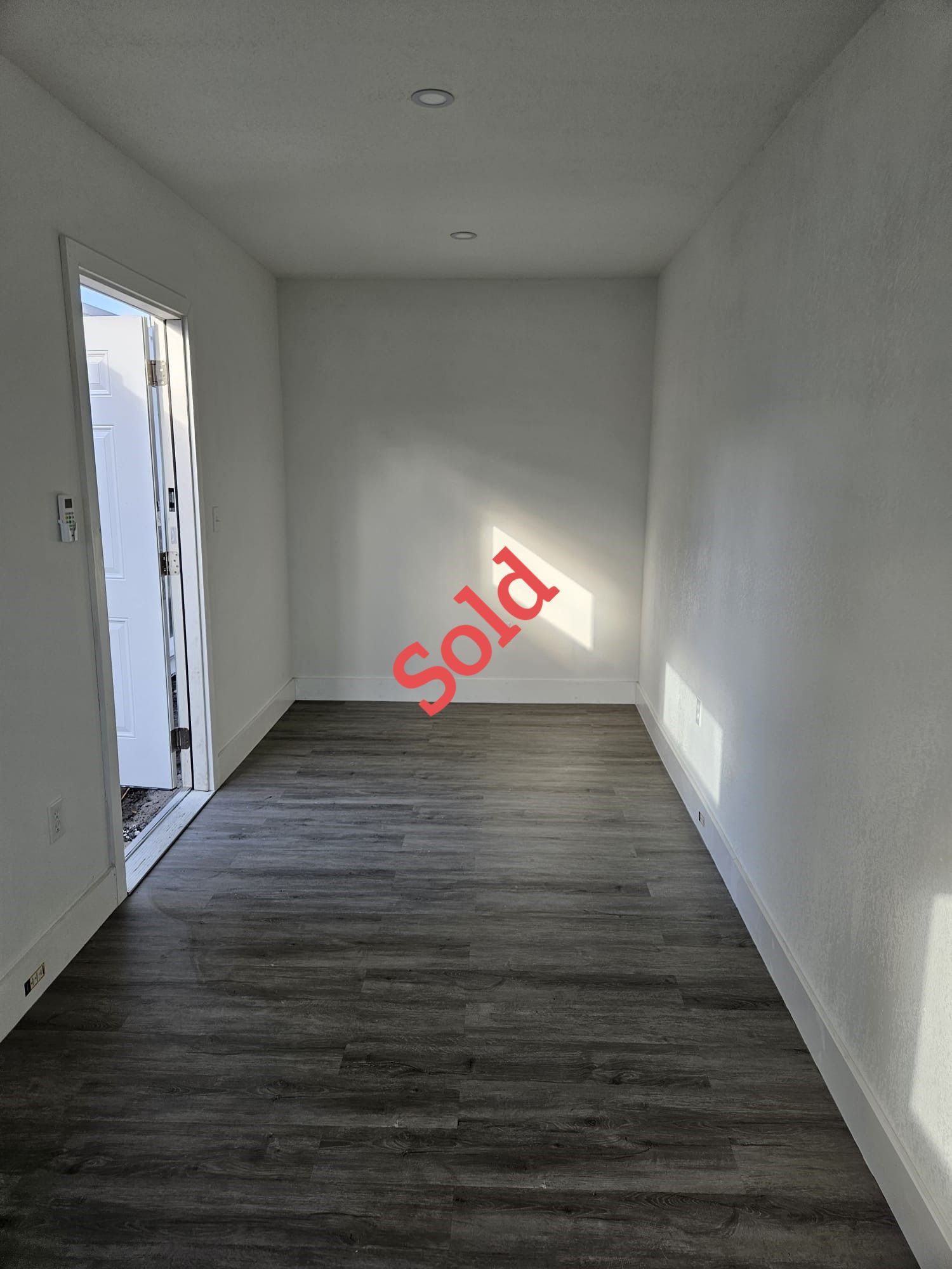 Empty room with dark gray wood floor, white walls, and a door on the left side, labeled 