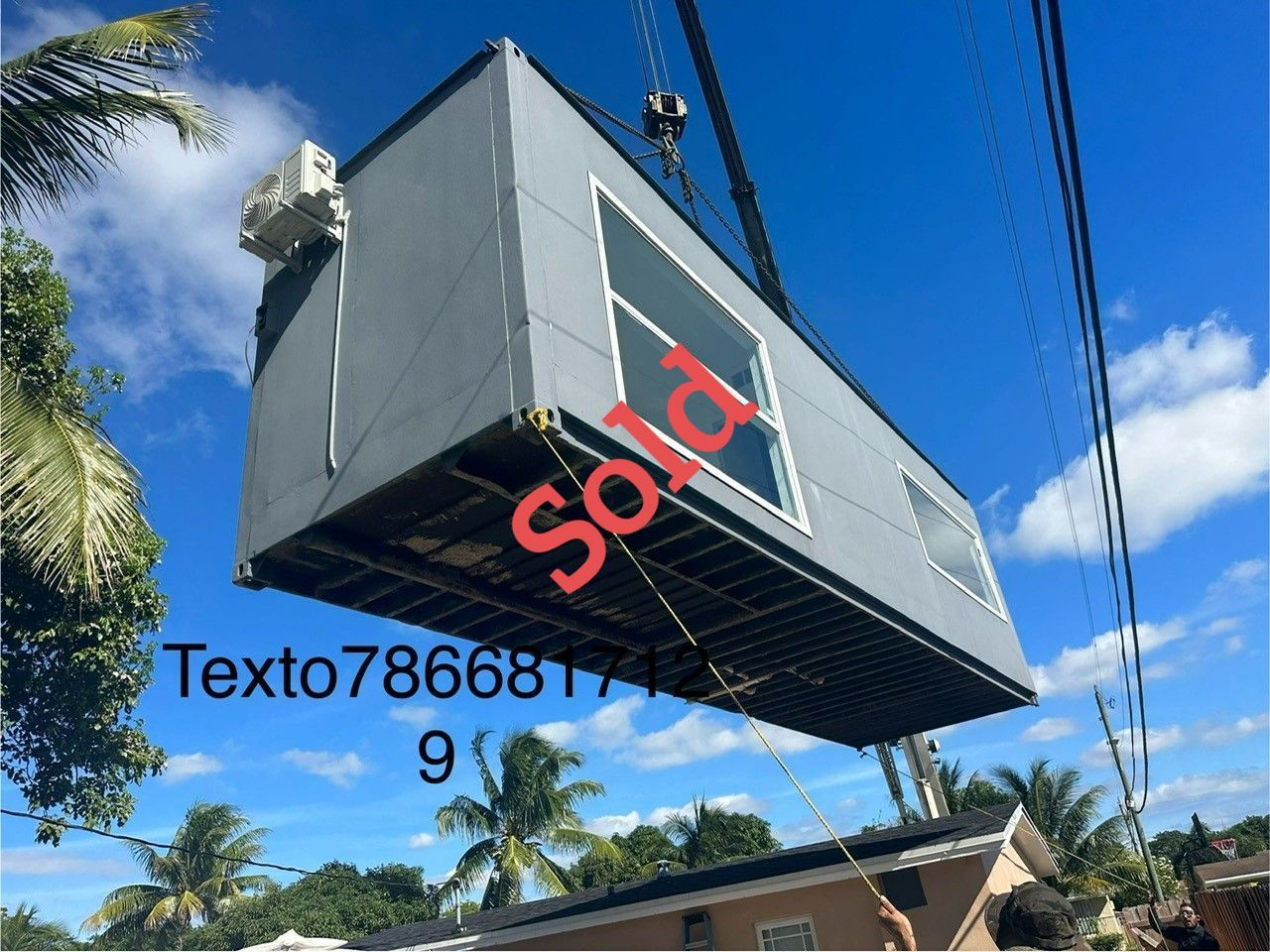 A modified shipping container, gray, is lifted by a crane. Sold sticker is on the container.