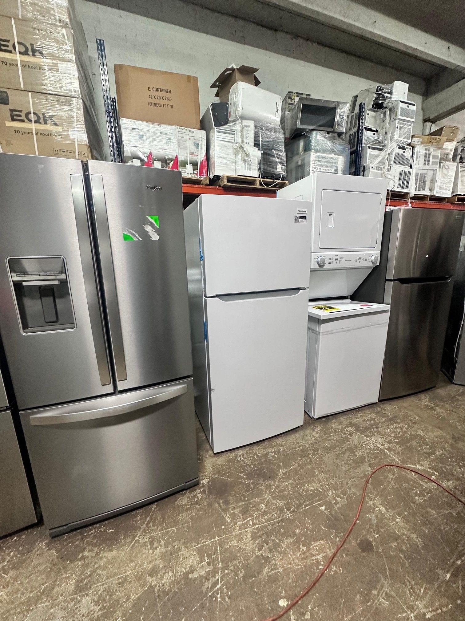 Refrigerators and appliances on display in a warehouse, including a stainless steel fridge and white fridge.