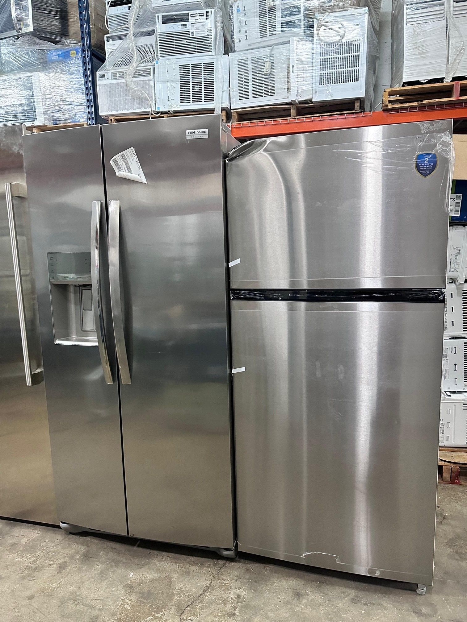 Two stainless steel refrigerators stand side-by-side in a warehouse. One is a side-by-side with a water dispenser.