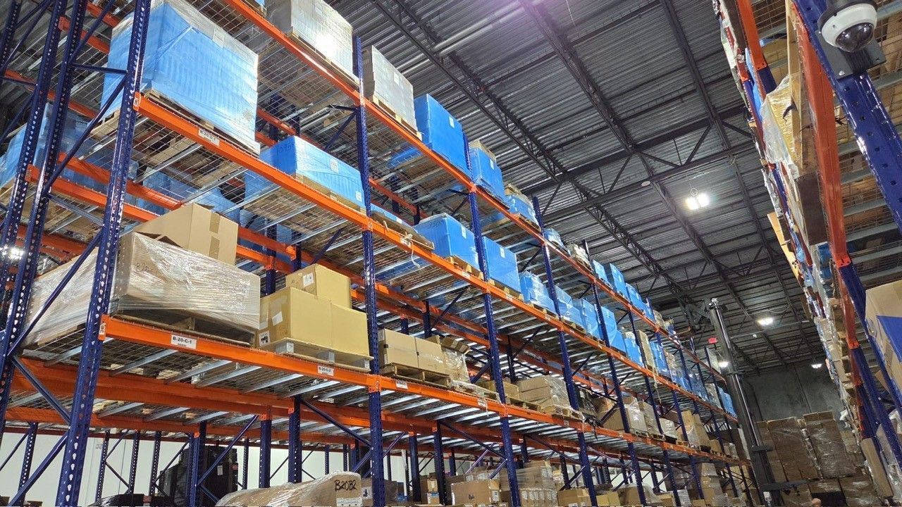 Warehouse interior with tall shelving filled with boxes and pallets, under a metal roof.
