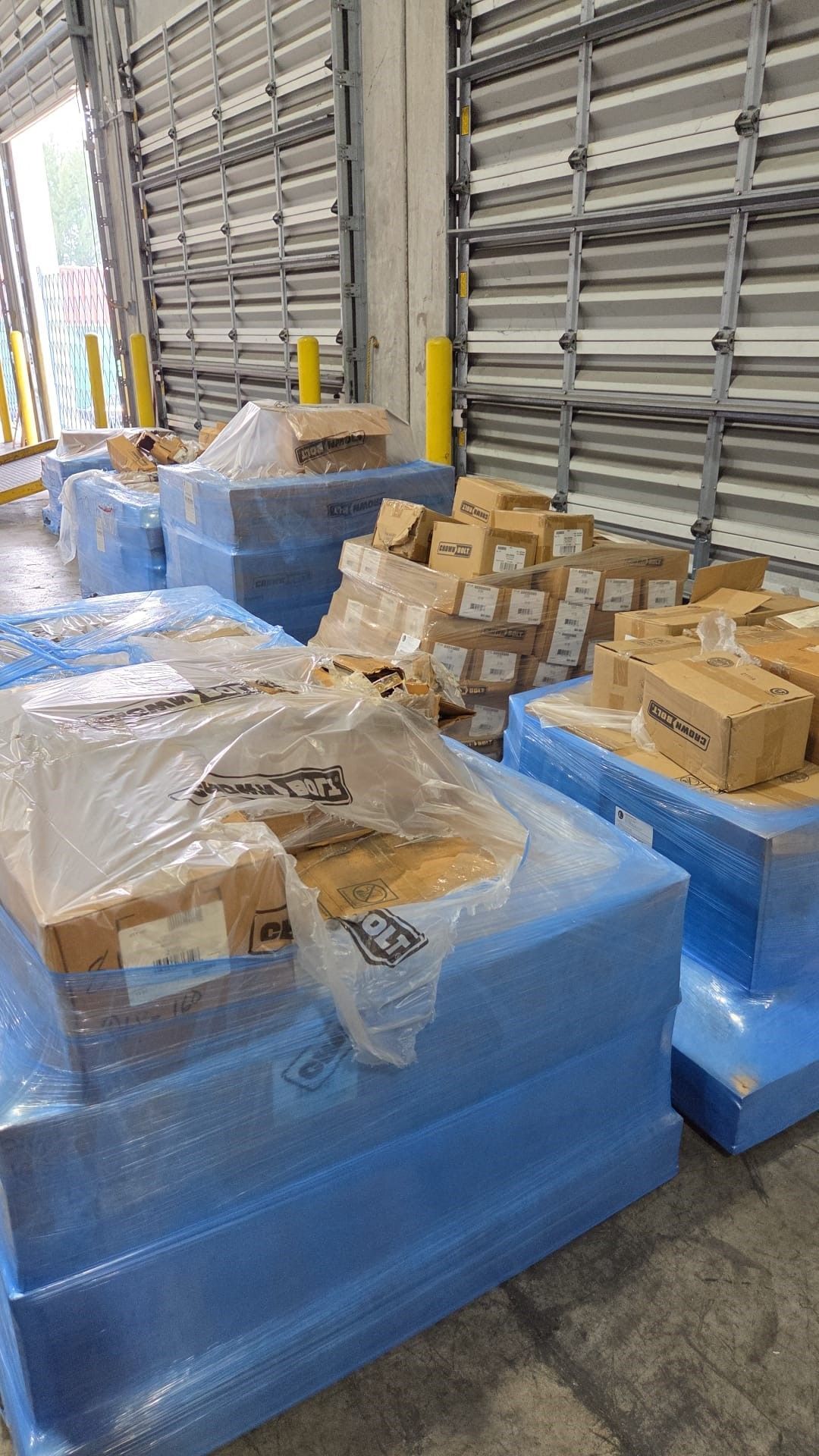 Pallets of blue-wrapped foam and cardboard boxes inside a warehouse, near a loading dock.