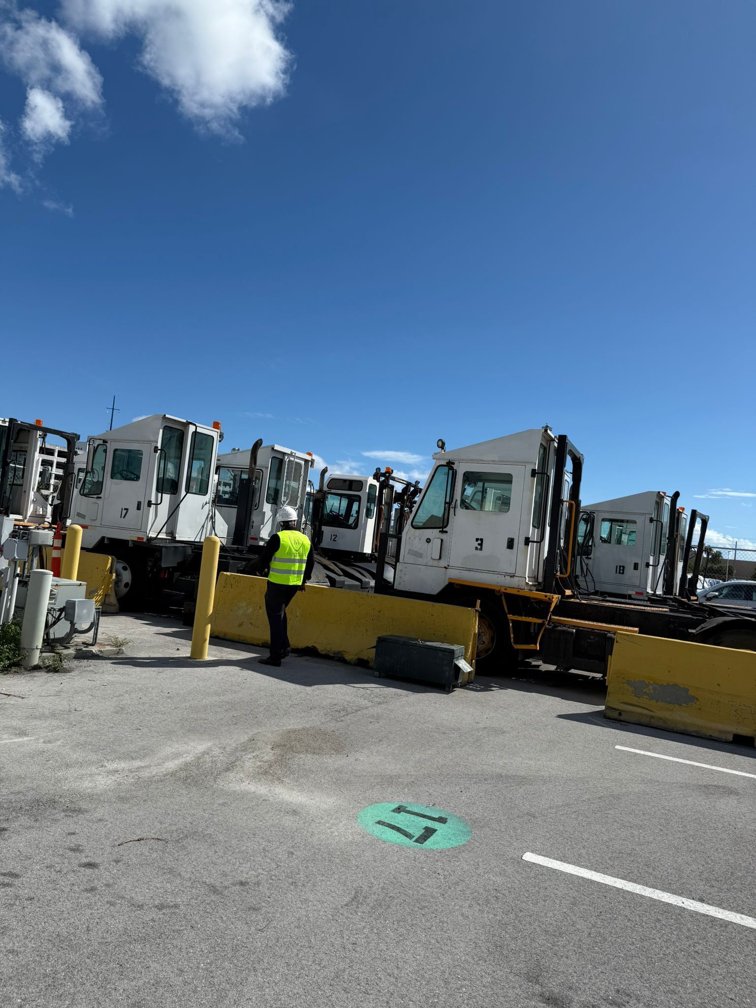 Person in a safety vest stands near parked white airport service vehicles on a paved lot under a blue sky.