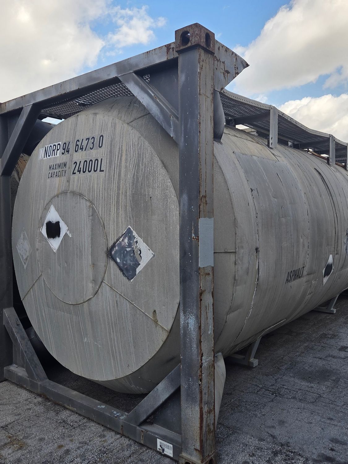 A large, cylindrical metal tank within a metal frame, featuring hazard labels and lettering. Outdoors, with a cloudy sky.