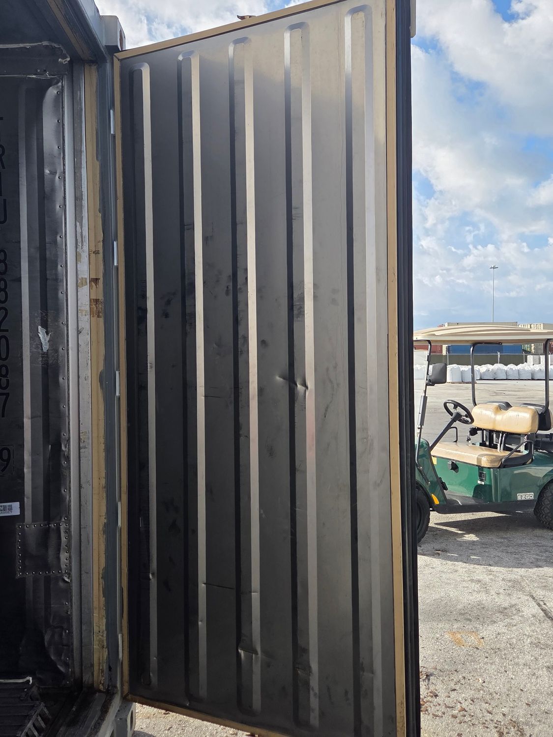 Open metal shipping container door, dented, revealing a golf cart and overcast sky.