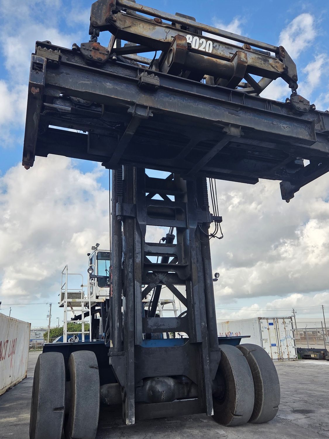 Large dark blue container handler, lifting apparatus extended against cloudy sky background.