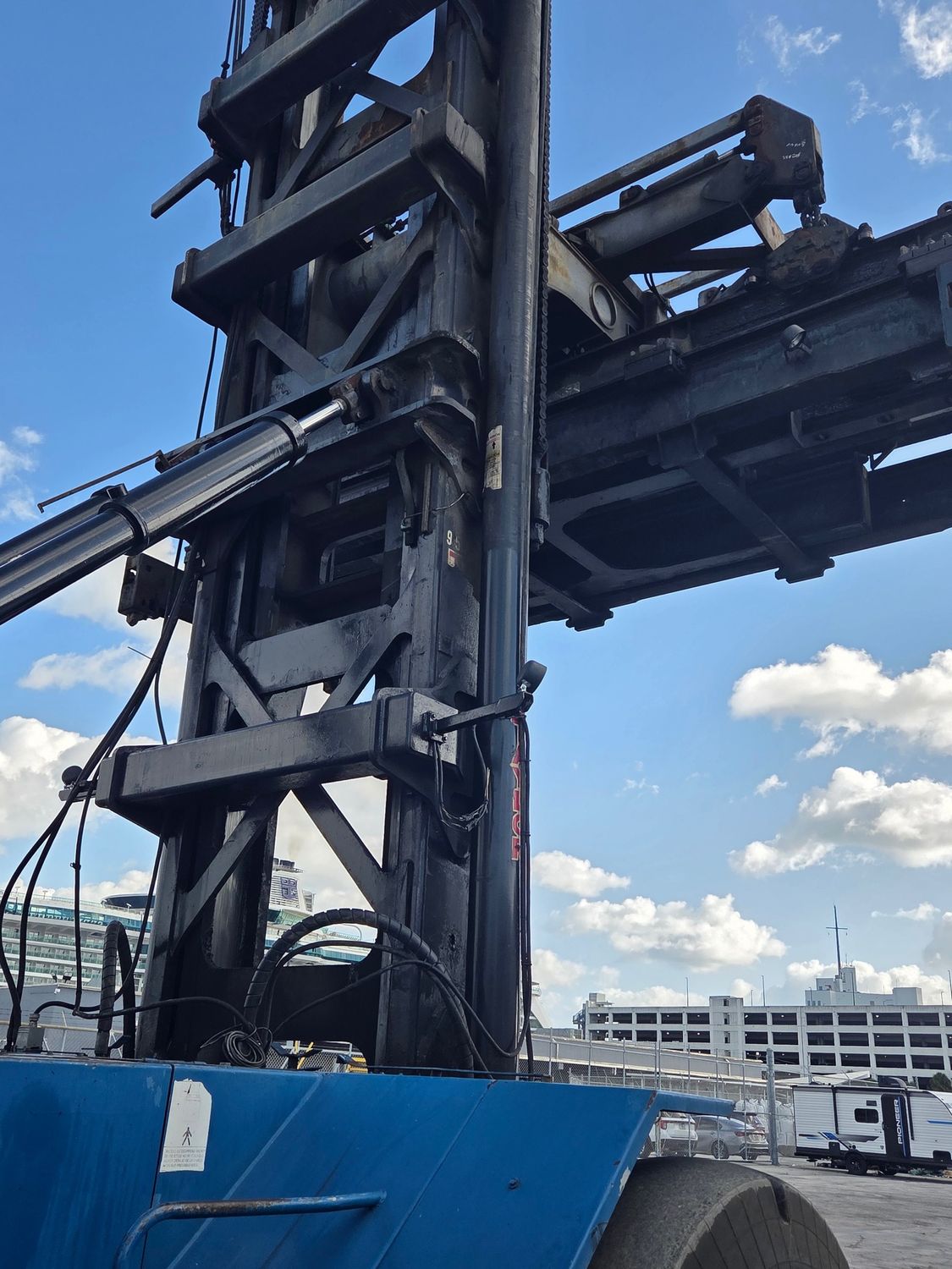 Blue and black industrial forklift against a blue sky, with a building and RVs visible in the distance.