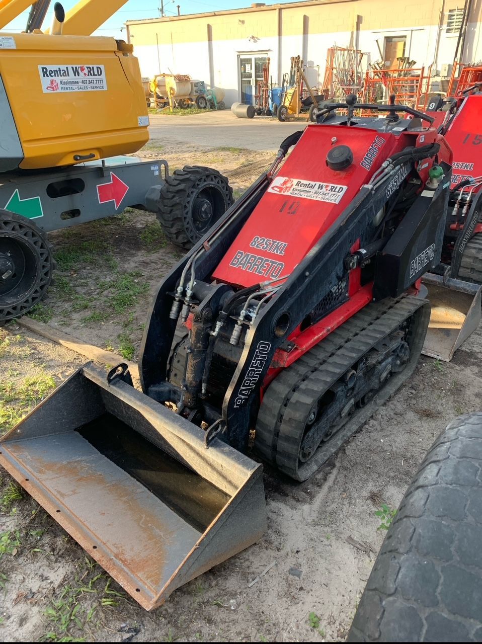 Red mini skid steer with bucket and tracks, parked on gravel next to yellow lift equipment.