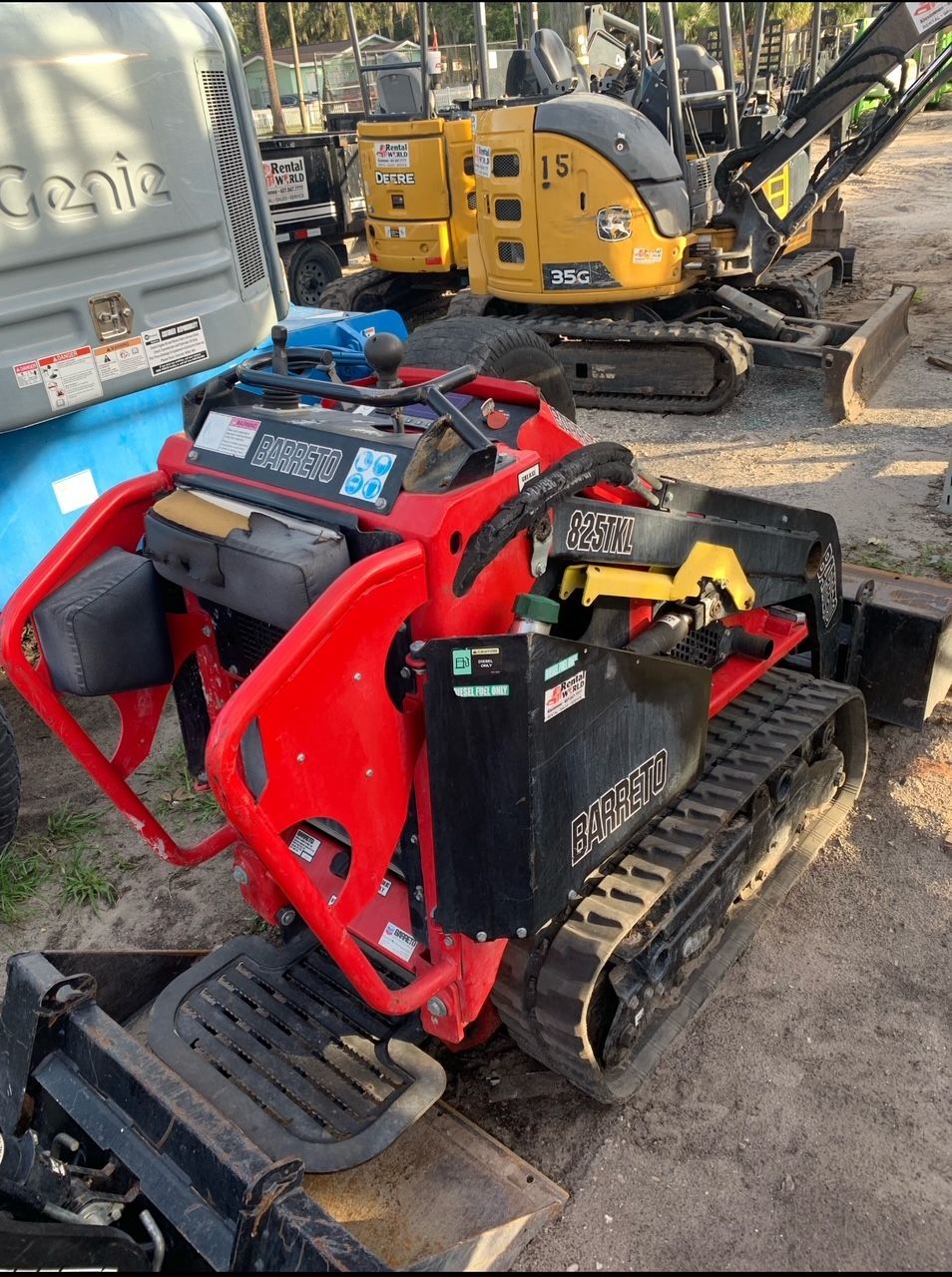 Red and black mini skid steer with tracks; other construction equipment in the background.