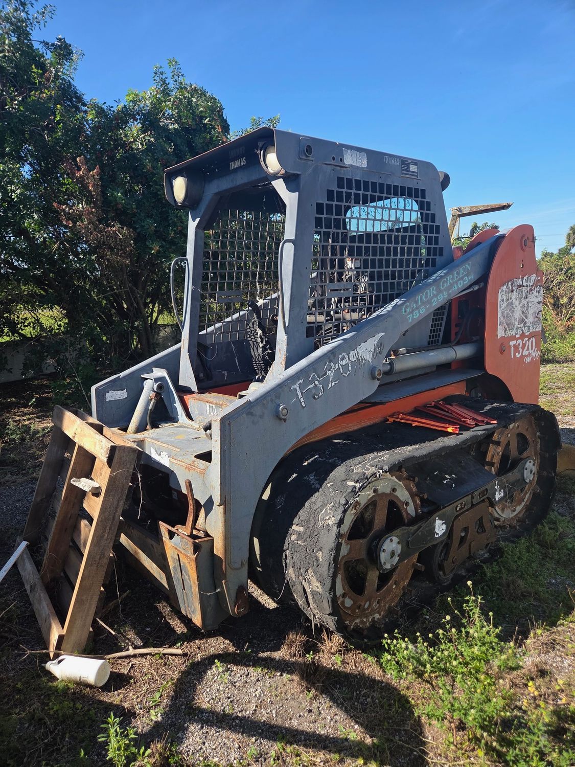 Gray and orange track skid steer on grass, in front of greenery and a blue sky.