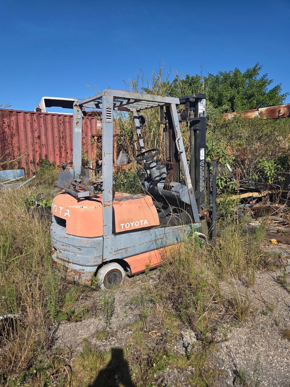 Abandoned, weathered Toyota forklift overgrown with weeds, orange and gray paint, outdoor setting.
