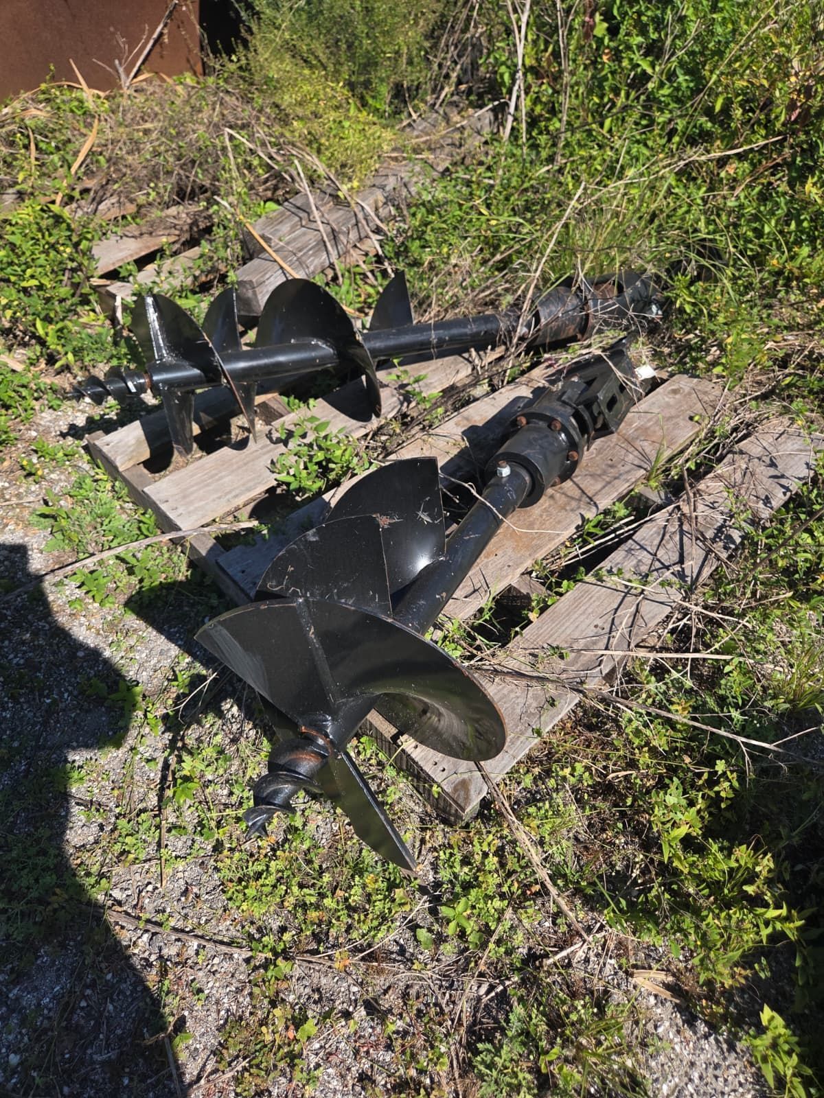 Two black augers and a boat propeller on wooden planks, outdoors.