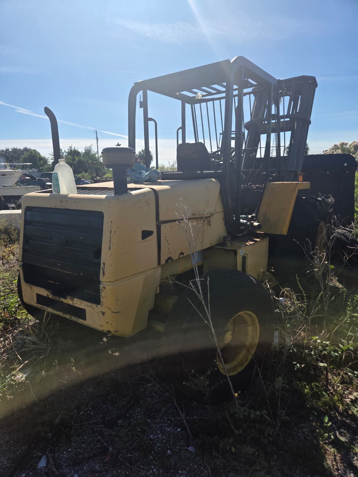 Yellow forklift parked outdoors with a black exhaust pipe and a protective cage, overgrown with weeds.