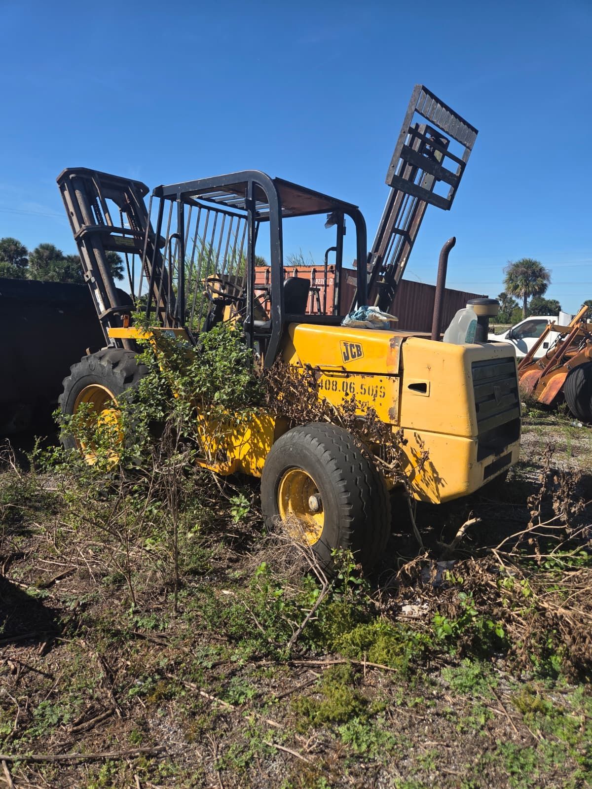 Yellow forklift in a field with raised forks, surrounded by overgrown vegetation.