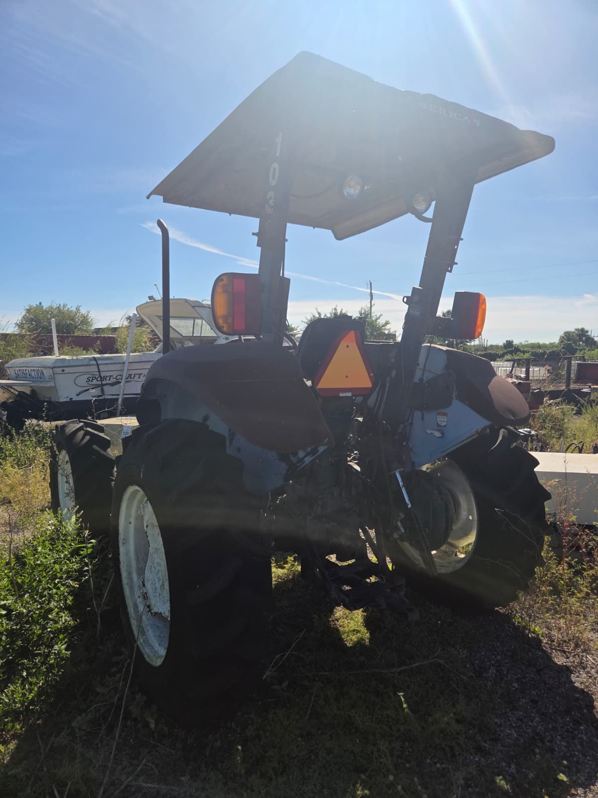 Rear view of a dark-colored tractor with a canopy, orange lights, and a white wheel, outdoors on a sunny day.