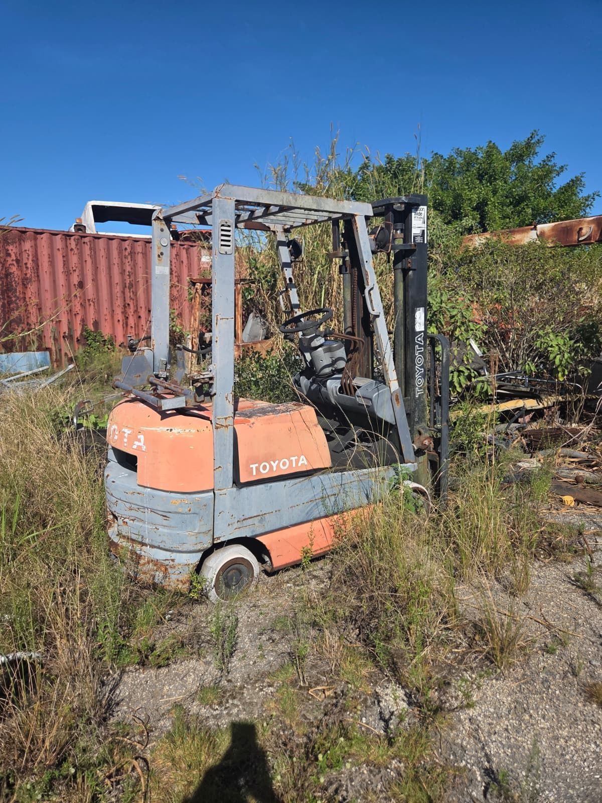 Old, weathered Toyota forklift in overgrown grass; orange and gray paint, visible in an outdoor setting.