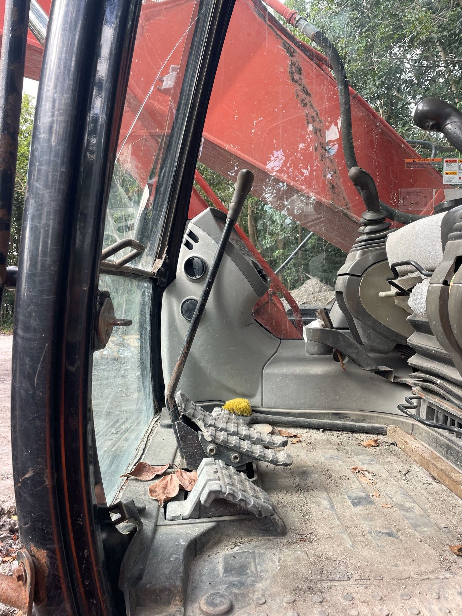 Interior of an excavator cab with controls, window, and exterior in the background. Cab is dusty.