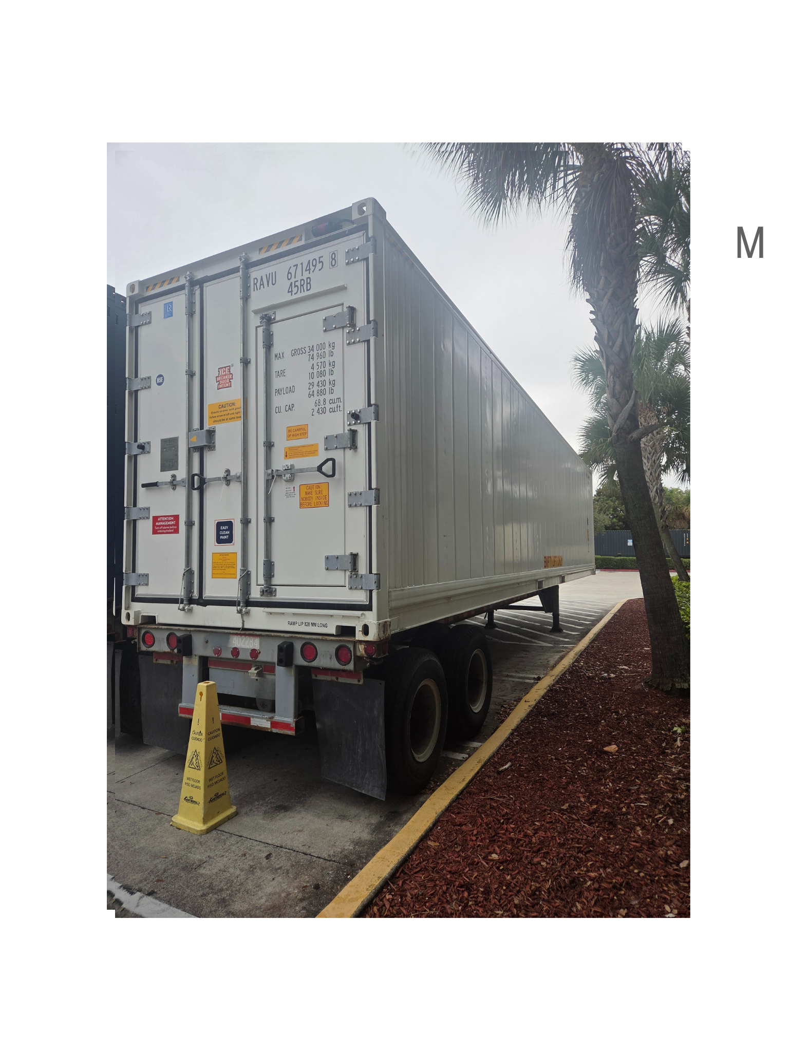 White refrigerated semi-trailer parked beside a building. Yellow traffic cone is placed behind the trailer.
