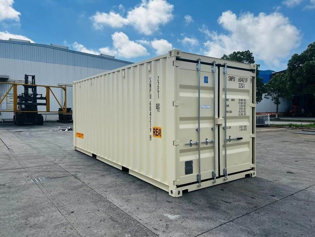 Beige shipping container on a concrete surface, with a warehouse and forklift in the background under a blue sky.