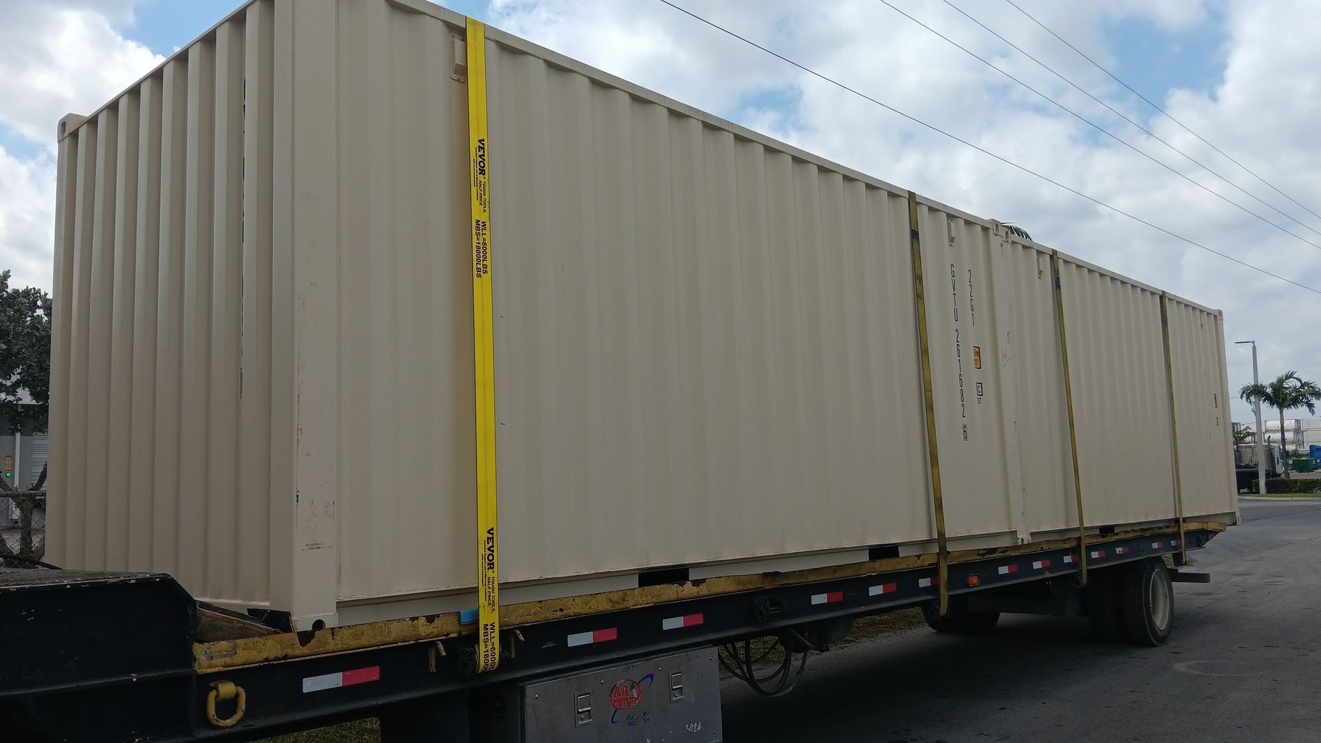 Tan shipping container secured on a flatbed trailer with yellow straps, outdoors.