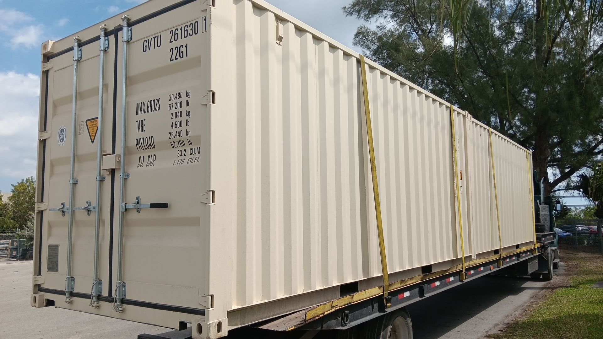Tan shipping container secured on a flatbed truck on a road. Bright, sunny day.