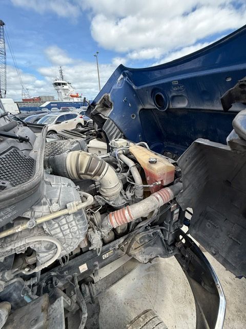 Truck engine bay with hood open; visible engine components, blue hood, and harbor background.