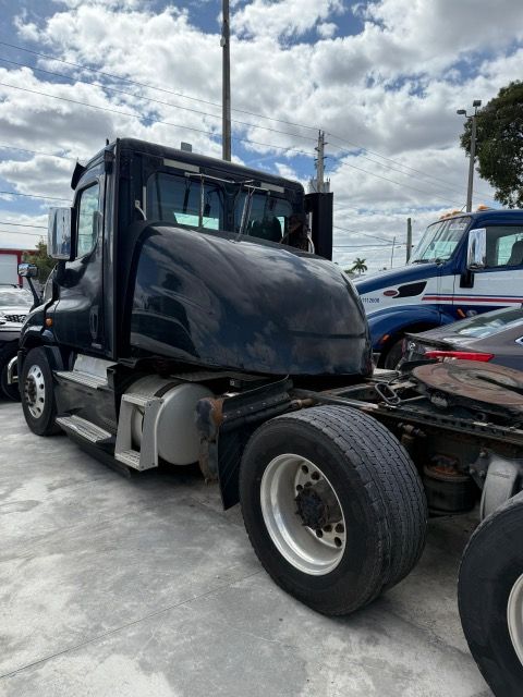 Black semi-truck parked outdoors on a gray surface, a partly cloudy sky overhead.