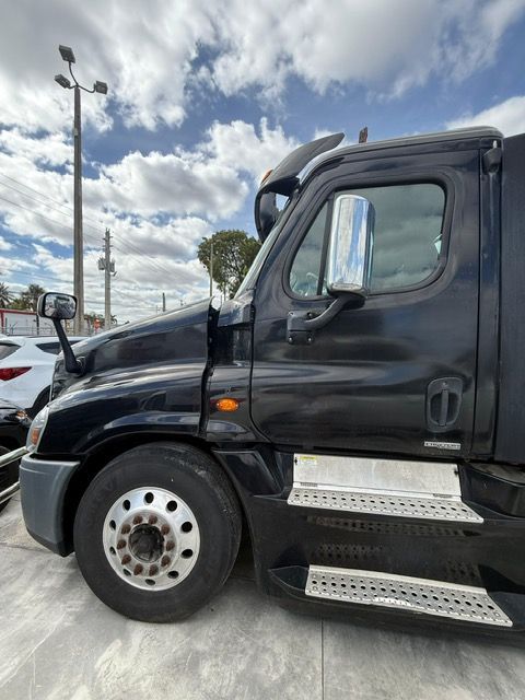 Black semi-truck, parked outdoors, under a cloudy sky. Side view of the cab with side mirrors and step.