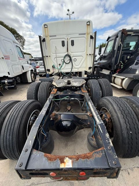 Rear view of a white semi-truck cab and chassis in a salvage yard, showing tires and exposed frame.