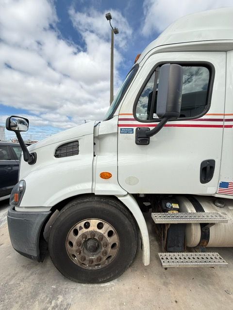 White semi-truck parked outdoors under a cloudy blue sky. Side view of the truck's cab and front tire.