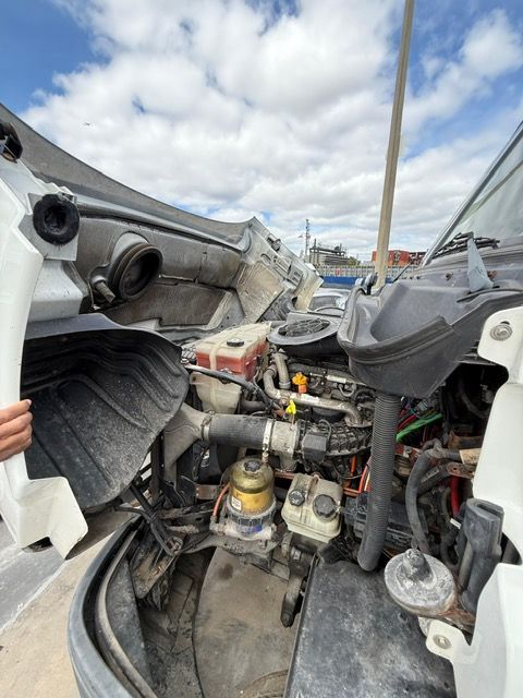 Open hood of a semi-truck revealing engine components, including hoses, filters, and reservoirs. Daytime outdoor setting.
