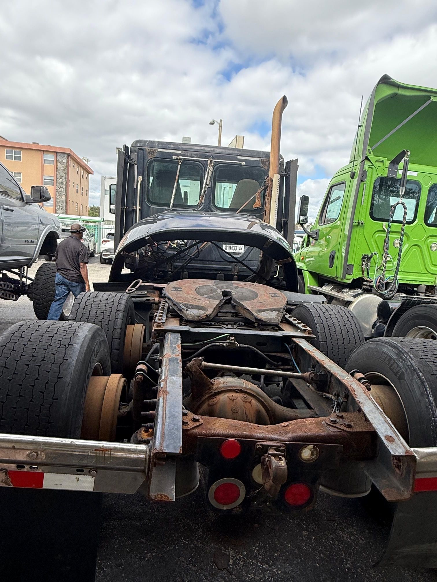 Rear view of a burned-out black semi-truck cab, exposing the chassis and fifth wheel; other trucks in background.