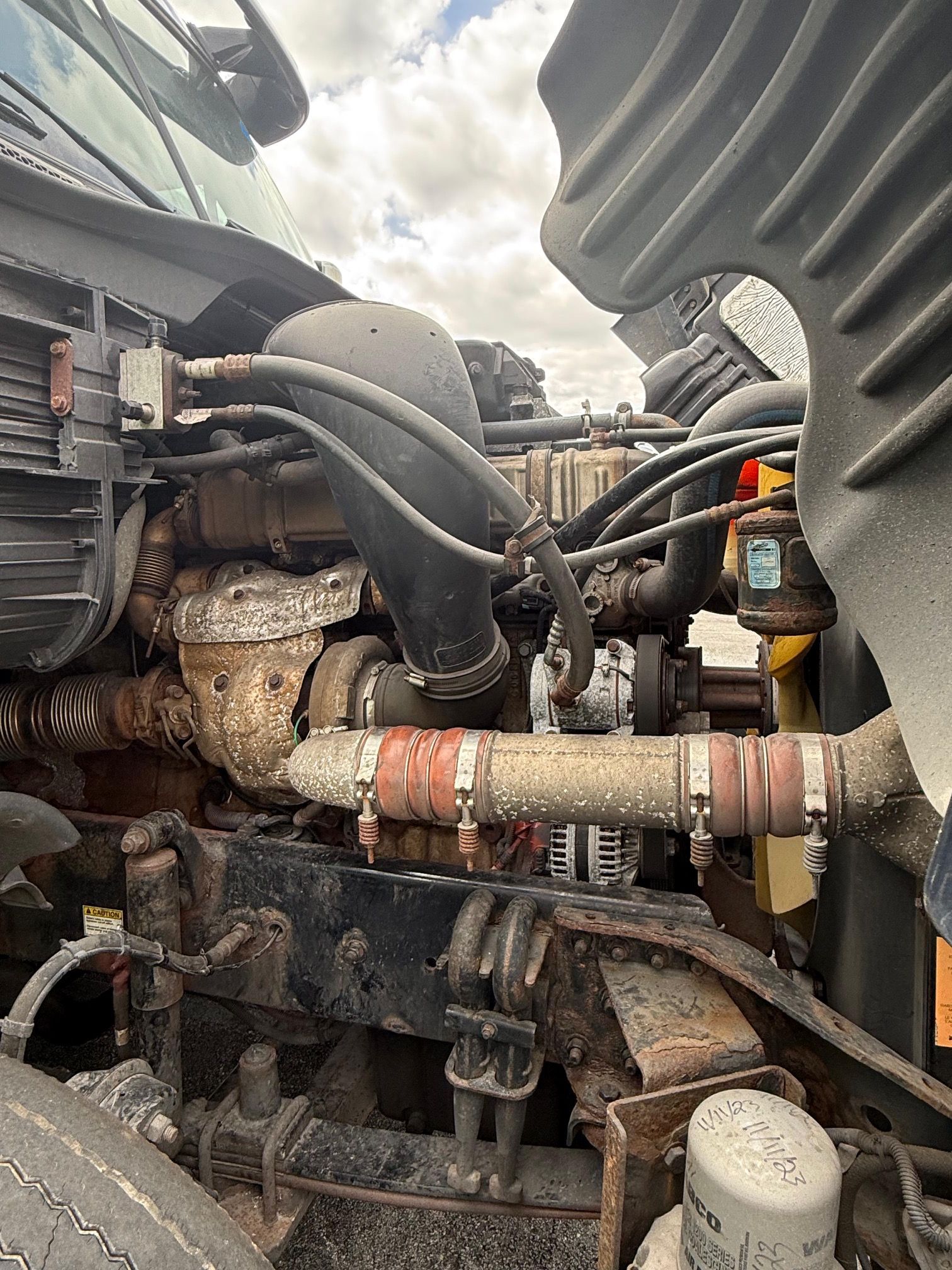Open engine bay of a truck with various rusted metal components and black tubing under a cloudy sky.