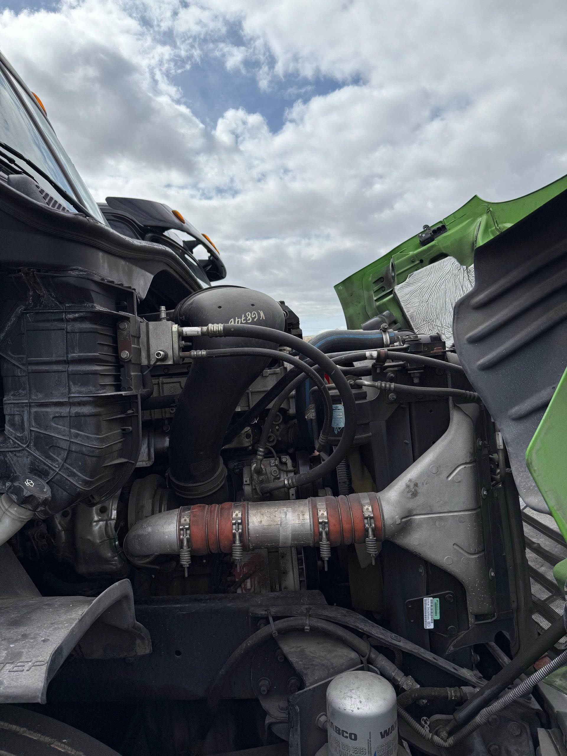 Open engine compartment of a green semi-truck with exposed machinery against a cloudy sky.