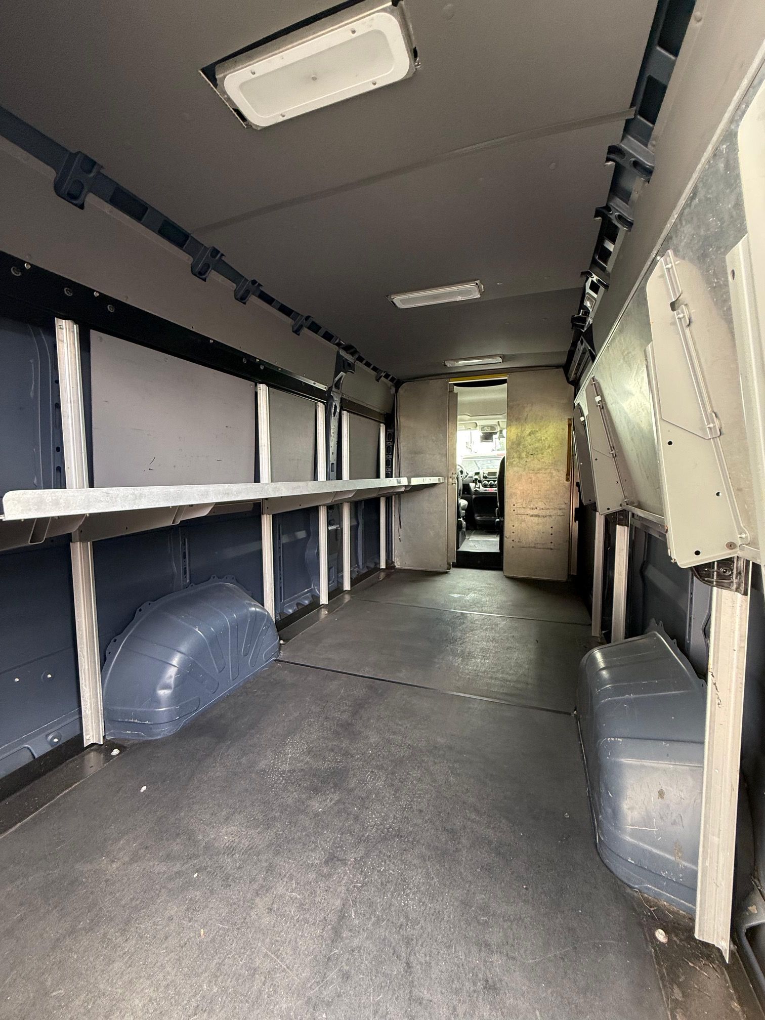 Interior of a cargo van with shelves along the walls and a view toward the front cabin. Grey walls, floors, and shelves.