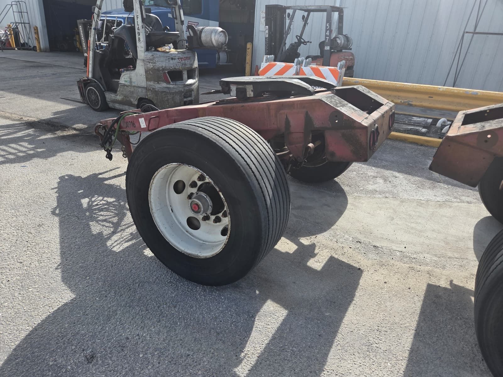A rusty trailer chassis with tires sits on a paved surface. A forklift and other equipment are in the background.