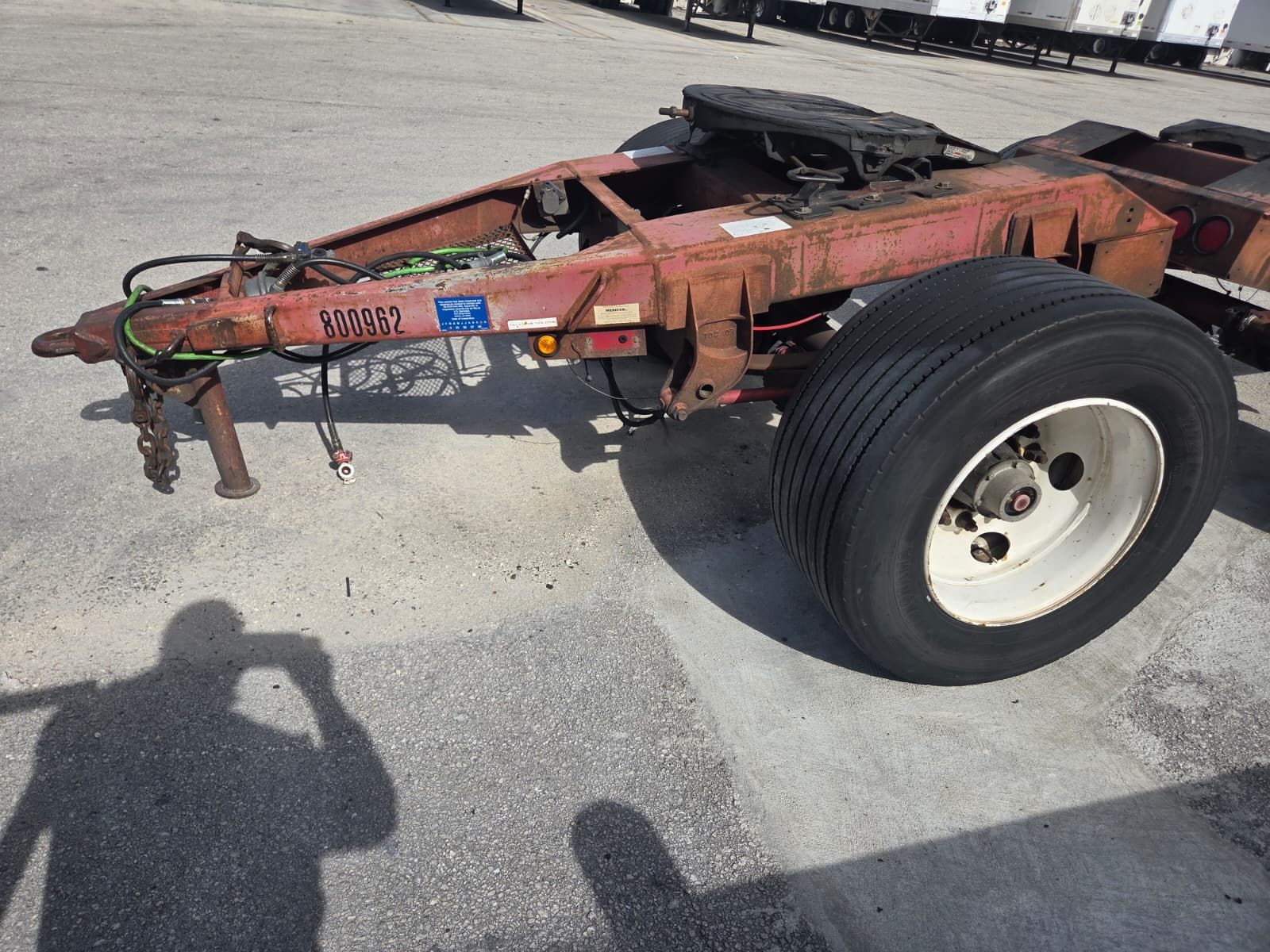 Red rusty semi-trailer chassis with a tire, parked on pavement, with electrical cords and hitch.