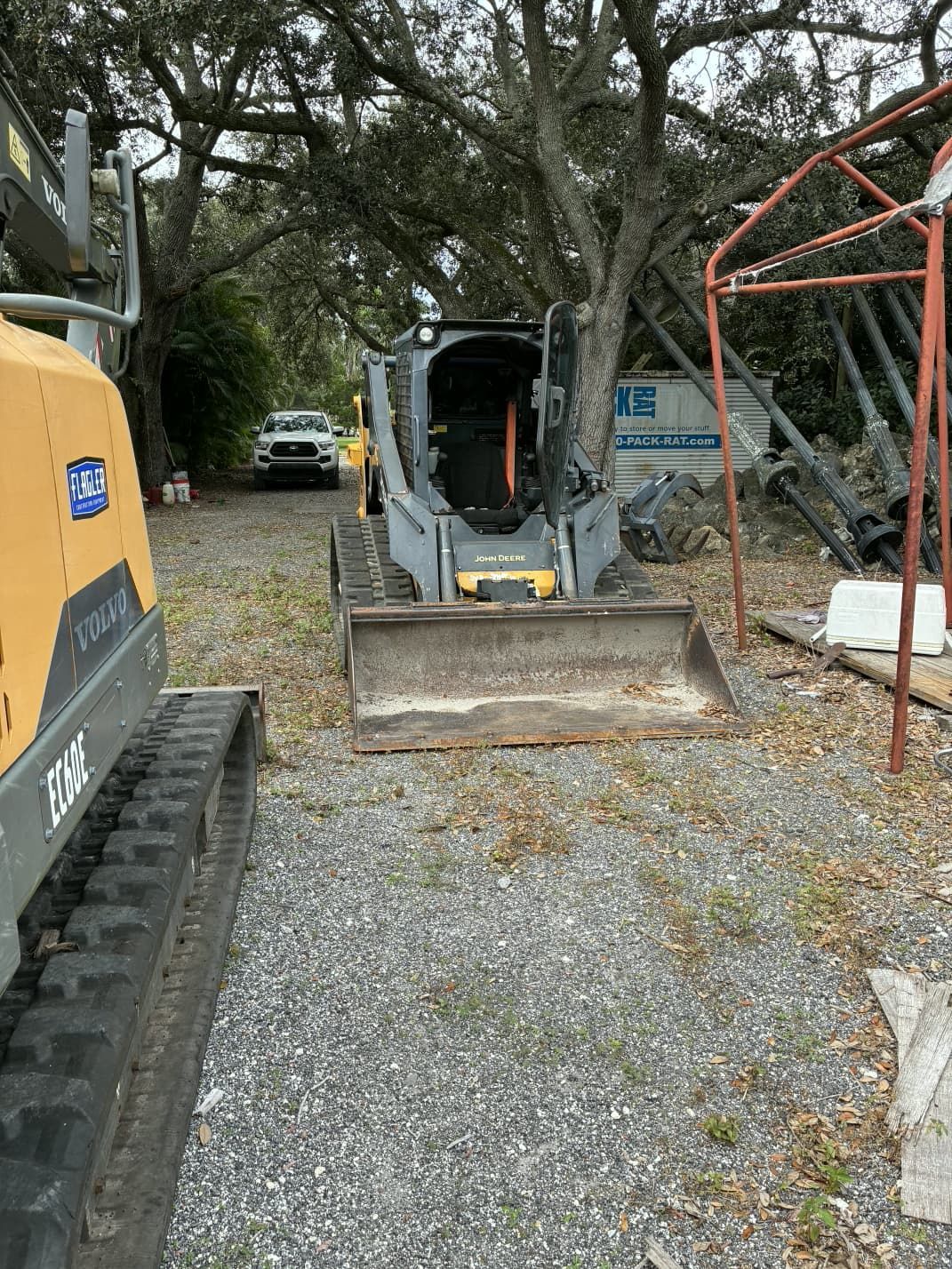 Skid steer loader parked near a tree and a small excavator on a gravel surface.