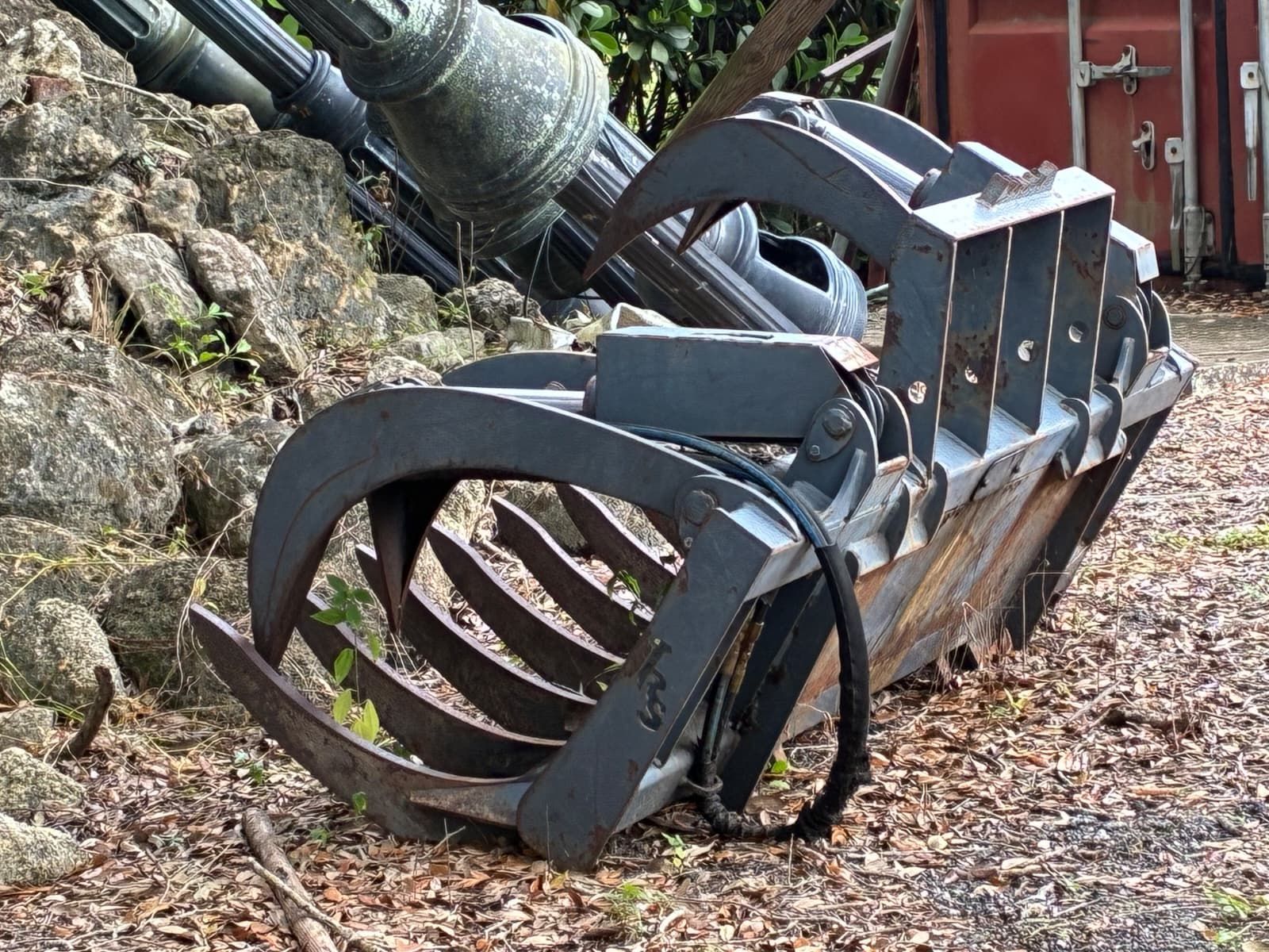 Heavy-duty grapple bucket, dark gray, resting on ground near stone and a container.