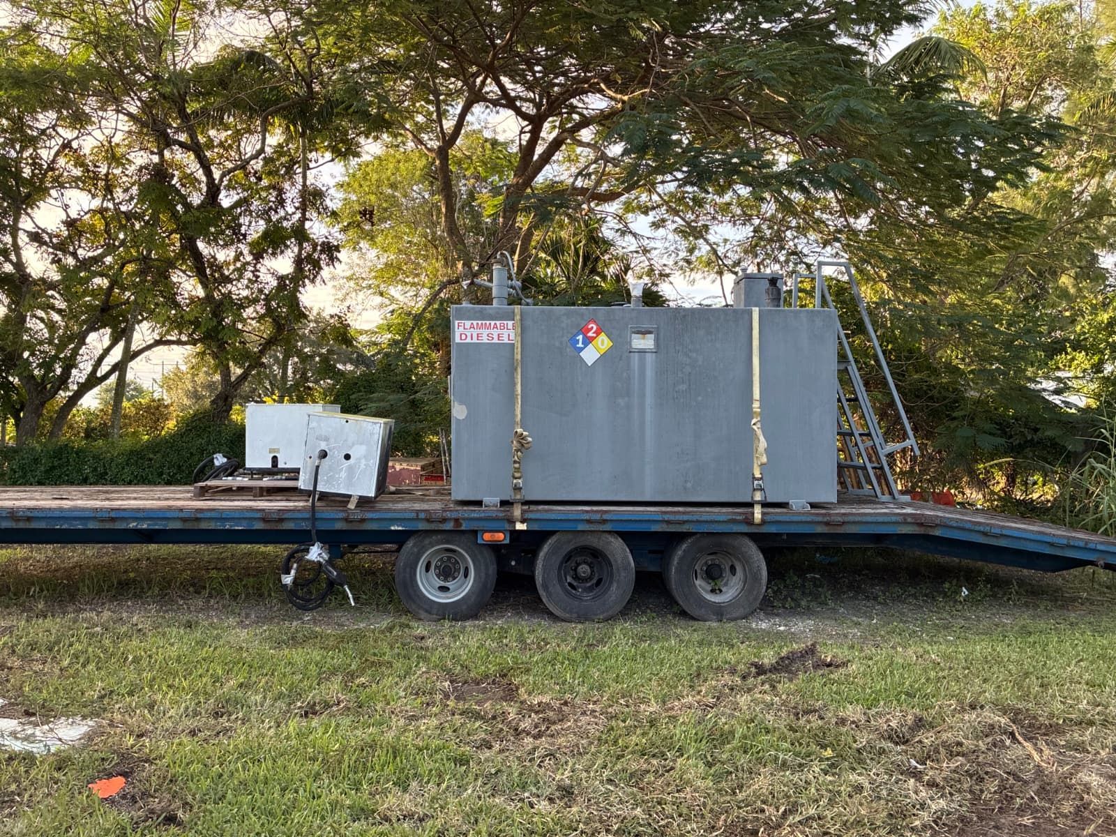Gray industrial tank on a flatbed trailer, secured with straps, in a grassy area with trees.