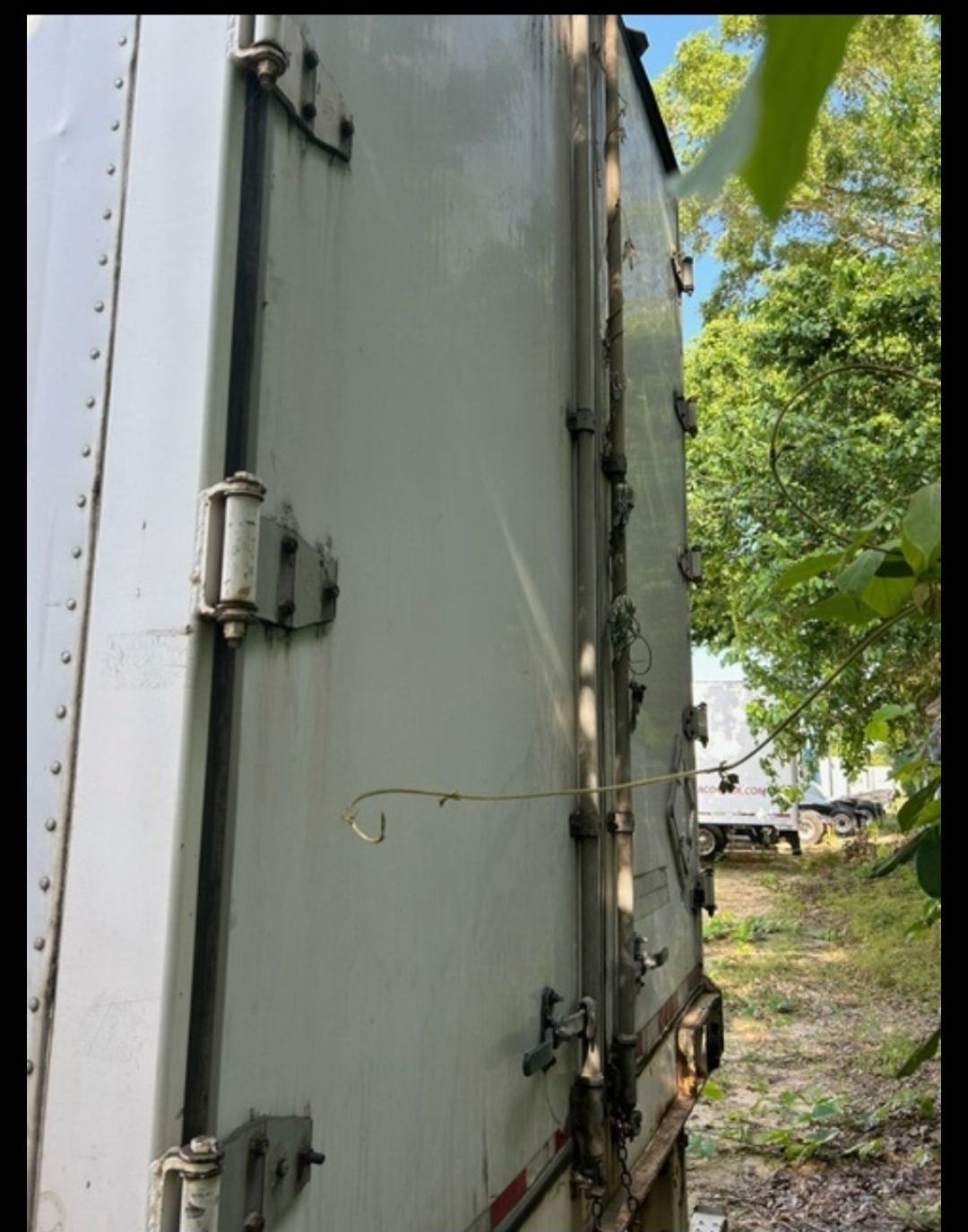 Side view of a white semi-trailer with latches, set outdoors near foliage and other vehicles.