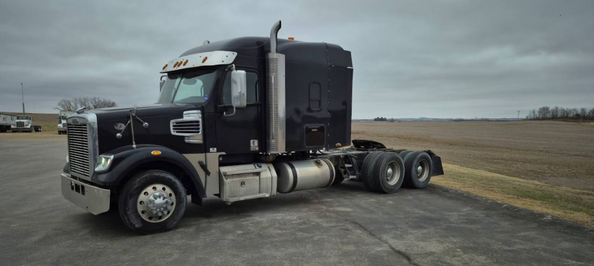 Black semi-truck parked on pavement in front of a harvested field under an overcast sky.