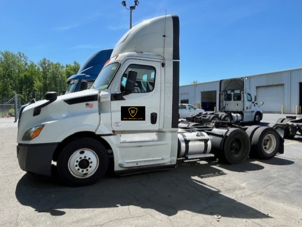 White semi-truck parked outdoors on a sunny day. Another truck visible in the background.